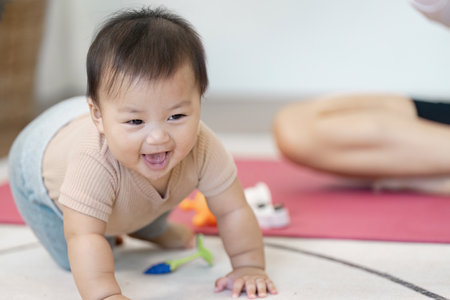 Joyful crawling baby exploring toys during playtime, with a multitasking mother nearby at homeの写真素材