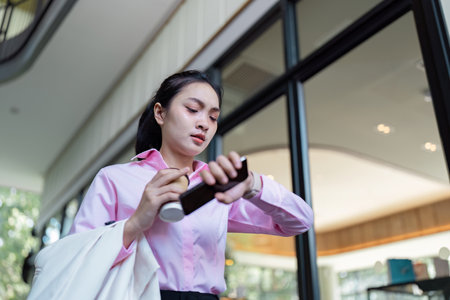 Focused Asian businesswoman checking her smartwatch while holding a coffee cup indoors, balancing work and daily routine.の写真素材