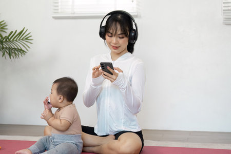Mother wearing headphones and using her phone while her baby plays beside her during a home workout...