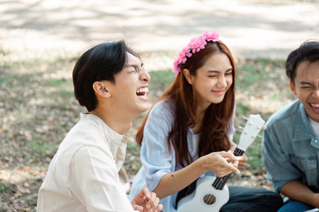 Diverse group of friends laughing and playing ukulele in a sunny park.の写真素材