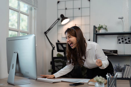Excited Asian female call center agent expressing enthusiasm after a successful customer call at a modern workspace.の写真素材