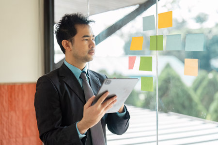 Thoughtful Asian businessman analyzing colorful sticky notes while holding a tablet in a modern office.の写真素材