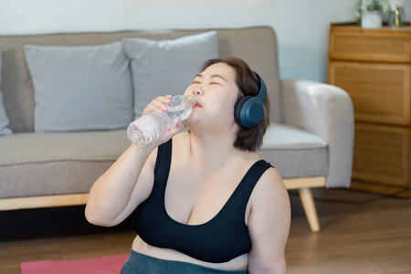 Hydrated woman enjoying water after workout while wearing headphones at home.の写真素材