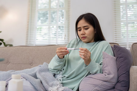 Woman checking her temperature with a thermometer while resting on the couch, managing her health.の写真素材