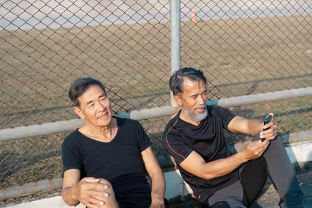 Content elderly men relaxing together and using a smartphone after a workout on the track. Emphasizing friendship, wellness, and active aging.の写真素材