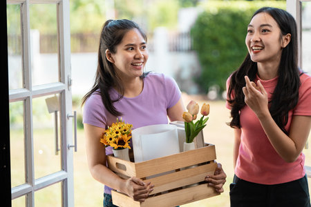Diverse female couple joyfully carrying flowers and unpacking boxes in their new home.