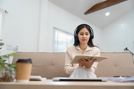 Young woman wearing headphones while reviewing notes on a couch, immersed in her study session.の写真素材