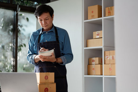 Business owner organizing packages and taking notes for online sales in a modern workspace.の写真素材