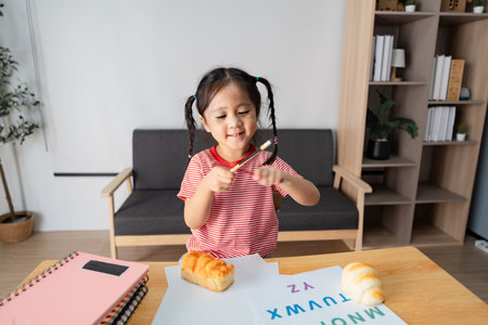 Cheerful girl holding a pencil and enjoying learning letters with snacks at homeの写真素材