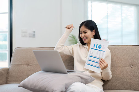 Excited young woman celebrating financial success while reviewing documents on a laptop at home.の写真素材