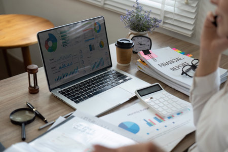 Financial analyst reviewing reports on a laptop while managing multiple financial documents.