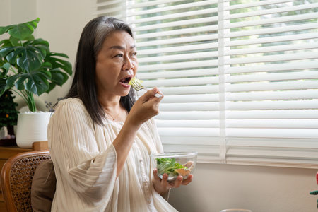 Wellness Habits and Nutritional Choices. An elderly woman relishing her salad in a cozy home setting.の写真素材