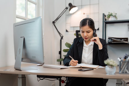 Focused Asian female call center agent taking notes while assisting customers in a modern workspace, emphasizing attention to detail.の写真素材