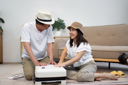 Elderly Asian couple smiling while packing a suitcase together, wearing hats and enjoying the travel preparation experience.の写真素材