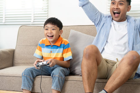 Joyful Family Moments and Gaming. A father and son share laughter during a gaming session in their...