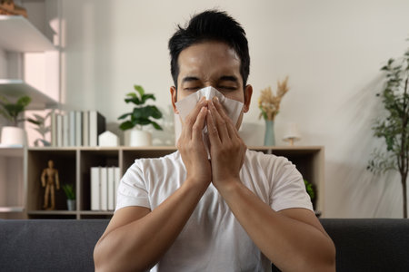 Allergy Symptoms. Man sneezing while wearing a mask indoors.の写真素材