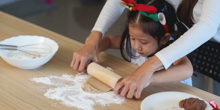 Christmas baking fun. A young girl rolls dough with her mother in a festive kitchen.