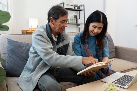 Elderly Man and Woman Sharing a Moment on the Couch with a Book and Laptop in a Cozy Living Room Settingの写真素材