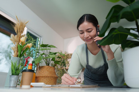 Business Call. Woman writing notes while talking on the phone in plant-filled space.の写真素材