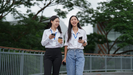 Two Asian businesswomen walking outdoors holding smartphones and coffee during work break