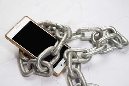 White smartphone with a large metal chain on a white background. Digital detoxification, dependence on technology, lack of concept of gadgets and devicesの写真素材