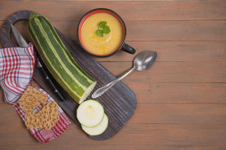 Autumn zucchini and pumpkin soup on a brown wooden background, sliced zucchini on a wooden cutting Board, grain crackers on a red napkin. Place for a copy spaceの写真素材