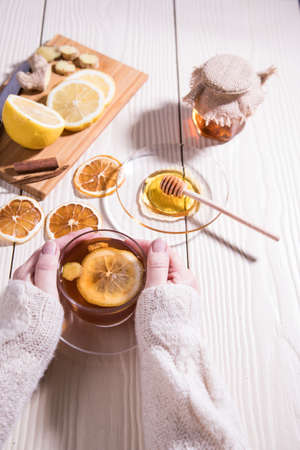 Women's hands in a white knitted sweater hold a Cup of warming tea with lemon, honey and ginger, white background.Close up.の写真素材