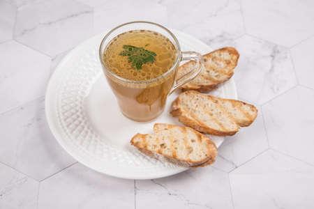 Homemade meat broth in a glass Cup on a white plate with crackers on a light background.の写真素材