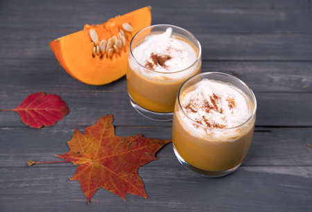 Two glasses of pumpkin latte with cream and cinnamon and a piece of sliced pumpkin on a dark wooden background. Autumn drinkの写真素材