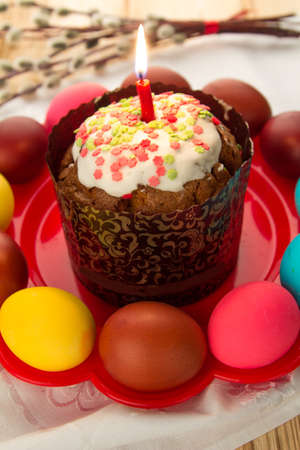 Close-up of a glazed Easter cake with a burning candle on a red plate with colored eggs.