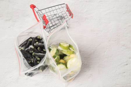 Bags of frozen zucchini and string beans stand on a light background next to a mini supermarket...