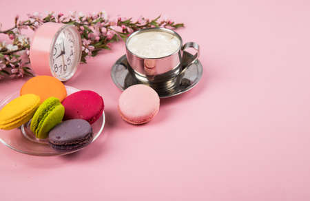 A cup of morning cappuccino with colorful macaroons cookies and a pink alarm clock on a pink paper background.の写真素材