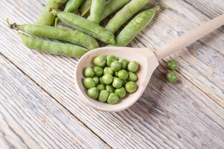 Top view of a wooden spoon with green peas and pods on a bright table.