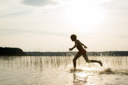 A slender guy child teenager runs along the water along the coast at sunset, splashing from the water.の写真素材