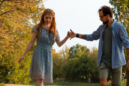 Young man helping girlfriend to ride longboard on sunny alley against green trees