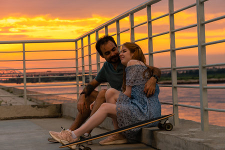 Young couple embracing while sitting on embankment against sunset skyの写真素材
