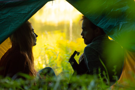Couple of travelers playing guitar while sitting in camping tent and looking each otherの写真素材