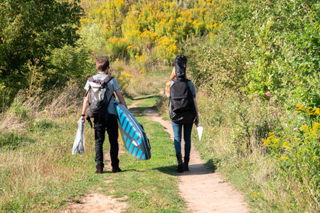 Unrecognizable travelers with backpack, guitar and sup board going through green countryside road