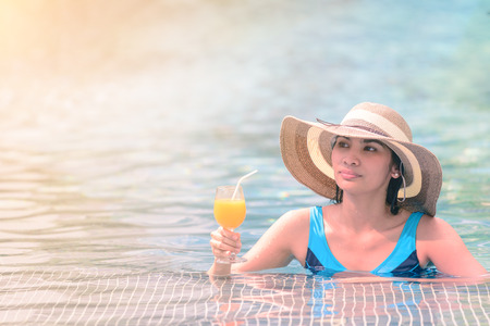 Beautiful woman in hat is holding orange juice glass at poolside in summer.