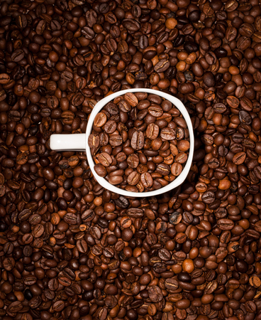 White cup full of coffee beans on Roasted Coffee Beans background
