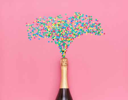 Creative photo of champagne bottle with colorful confetti on pink  background. Flat lay of christmas, anniversary, new year celebration conceptの写真素材