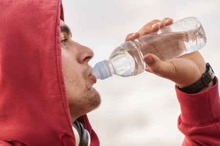 Young handsome man waring his red hoos on and drinking water from the small bottle and wearing headpones on his around his neck while looking it the sky. Bright shy is in backgroundの写真素材