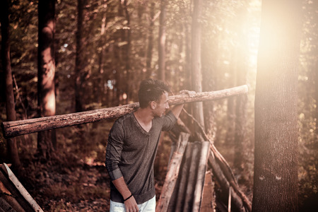 Young handsome man carrying heavy log on his shoulder as a survivor at the forest. He is looking aside over his shoulder while the forest is in background and sun rising from behind.の写真素材