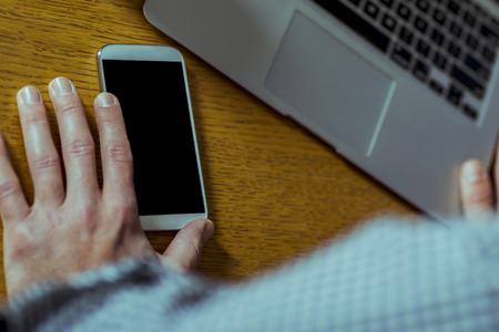 Close up of a modern smart phone and laptop next each other and hand leaning against the desk.