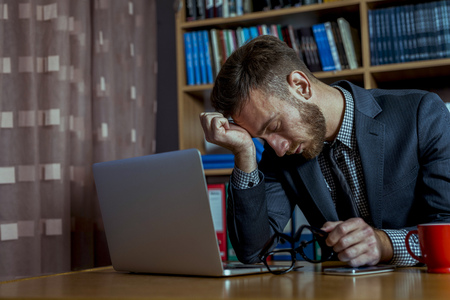 Handsome man using smart phone and computer at his office. Bookshelf is in background and laptop in front of a businessman. Cup of coffee is on the desk besides computer.の写真素材