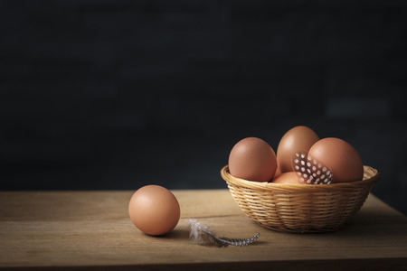 brown eggs in a wicker basket whit feathers on a wooden board set against a black backgroundの写真素材