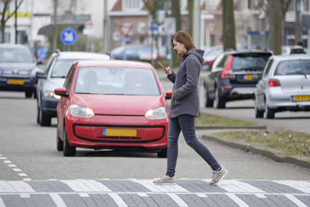 young woman walks on the crosswalk and checks her smartphone for messages and does not pay any attention to traffic.の写真素材