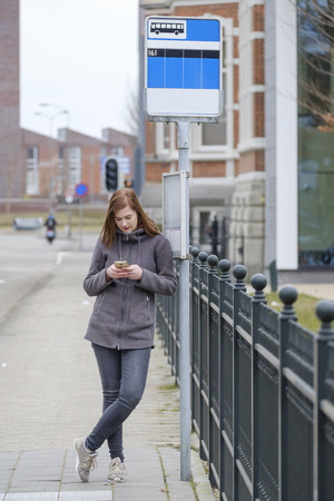 young woman stands at a bus stop and look while waiting boredom on her phoneの写真素材