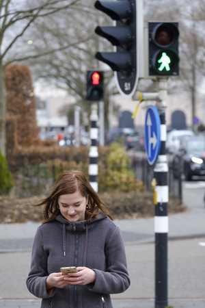 Young woman crossing the street, while looking at her cell phone but do not mind the traffic lightsの写真素材