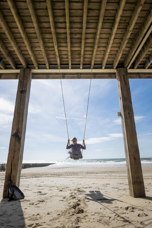 woman sitting in a decorated swing on the beach overlooking the sea on a sunny day with a blue skyの写真素材